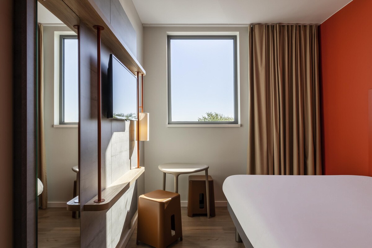 Habitación de hotel con cama, televisor montado en un panel de madera, una mesita, un taburete y una ventana con cortinas.