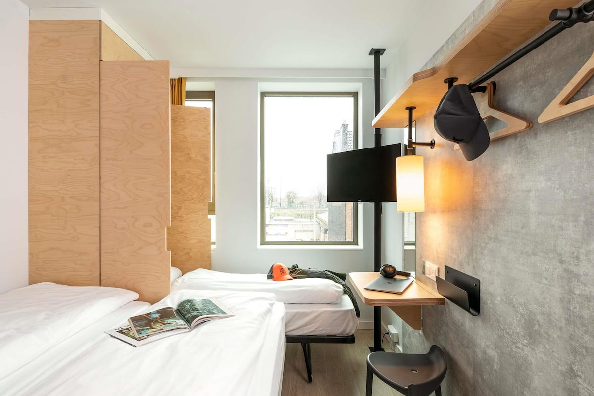 Habitació d'hotel amb dos llits, una taula de treball amb un ordinador portàtil, una cadira, un televisor i una finestra amb vista a edificis.