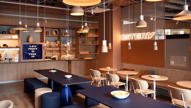 Una cafeteria moderna amb diverses zones d'ambientació, incloent taules i bancs, i un mostrador visible amb un cartell de neó.