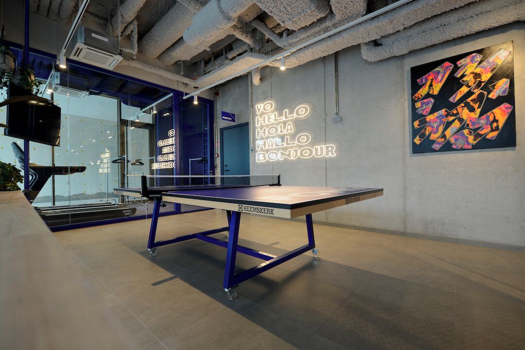 A ping pong table in a modern indoor space with a neon sign on the wall and a colorful abstract painting.