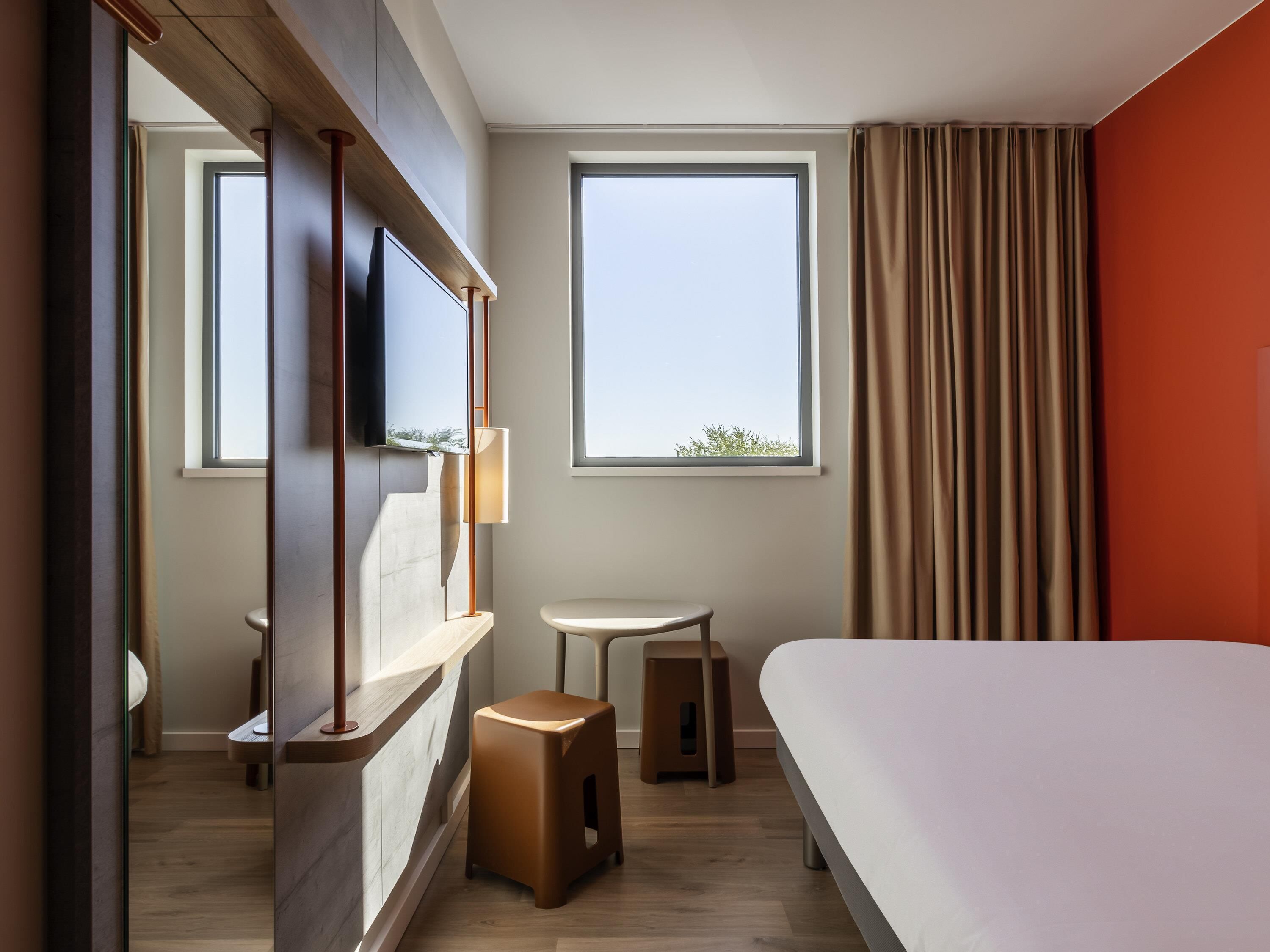 A hotel room with a bed, a TV mounted on a wooden panel, a small table, a stool, and a window with curtains.