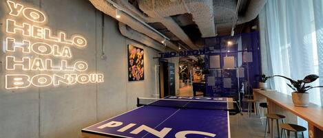 Una mesa de ping pong en un espacio interior moderno con techo de hormigón y un letrero de neón que dice "YO HELLO HOLA BONJOUR" en la pared.