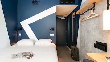 A modern hotel room with a bed, a desk, a chair, a hanging light fixture, and a coat rack.
