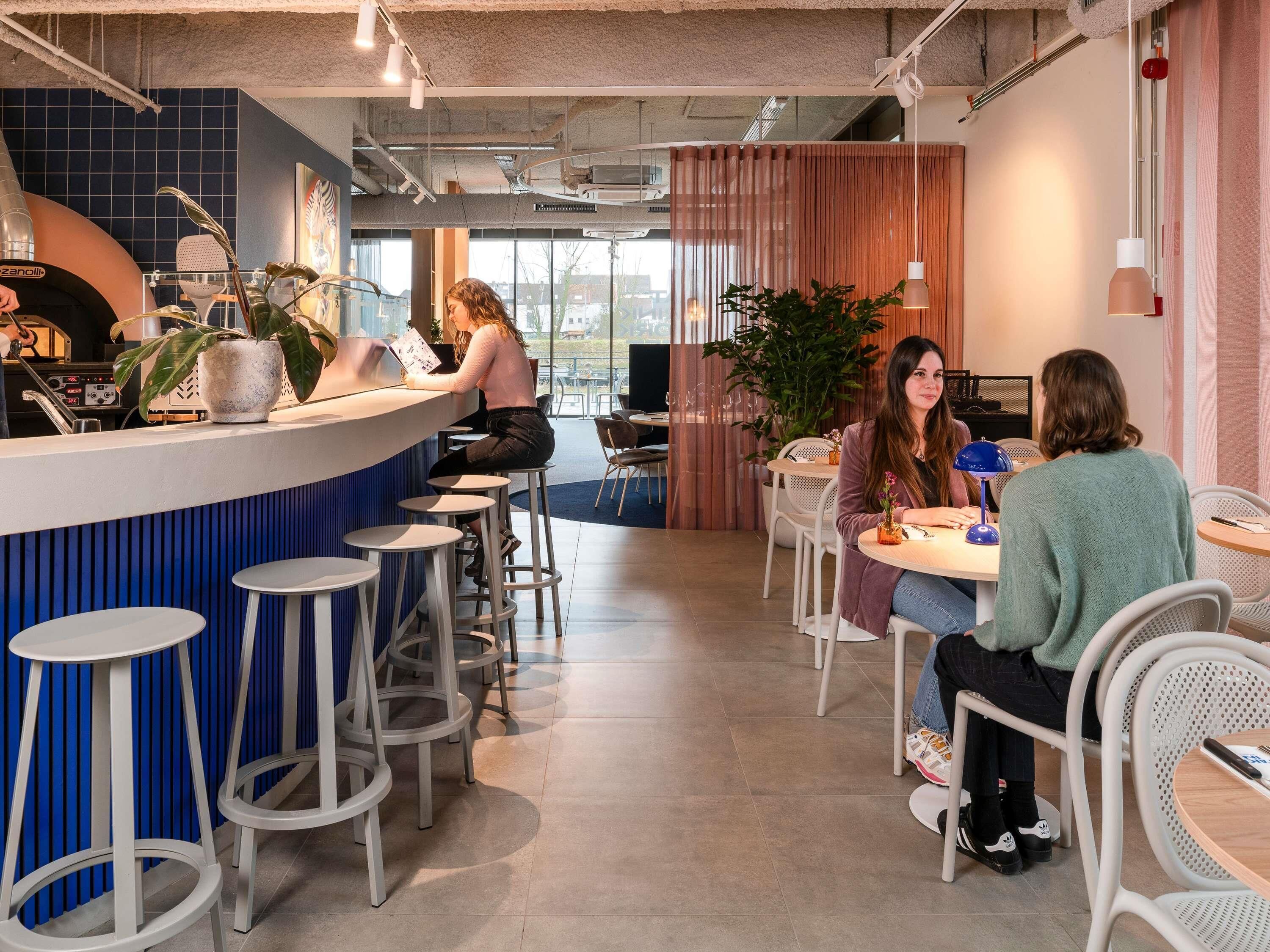 Interior de una cafetería moderna con mostrador, taburetes, mesas y plantas en macetas.
