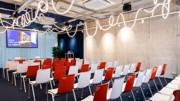 A conference room with rows of chairs, a projector screen, and a large plant.