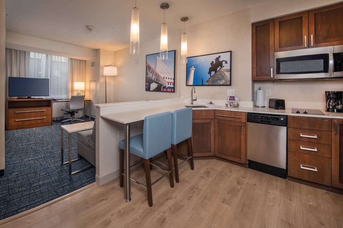 Una habitación de hotel moderna con kitchenette, una zona de comedor con sillas azules, un televisor de pantalla plana, un escritorio y un amplio ventanal con cortinas.