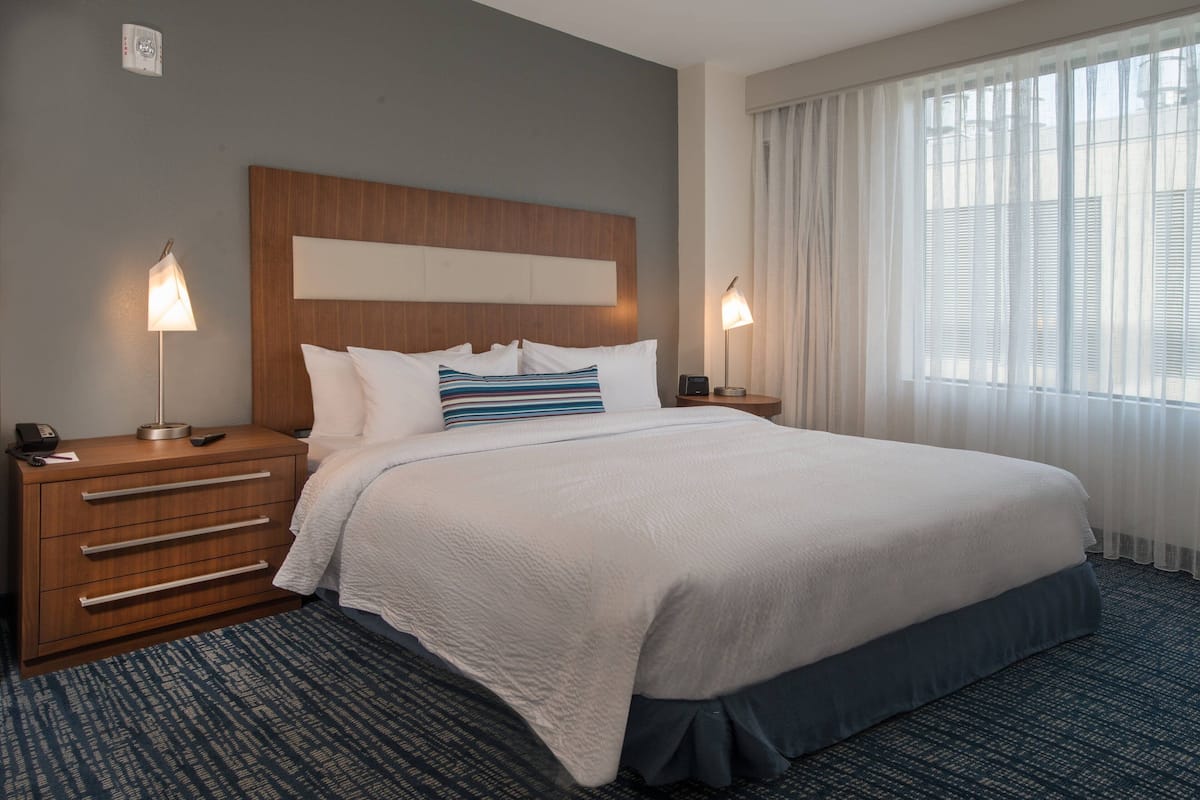 Habitación de hotel con una cama grande, cabecera de madera, dos mesitas de noche con lámparas, un aparador y una alfombra estampada.