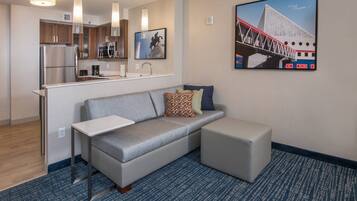A modern hotel room with a kitchenette, a gray sofa with patterned pillows, a small table, and a blue carpet.