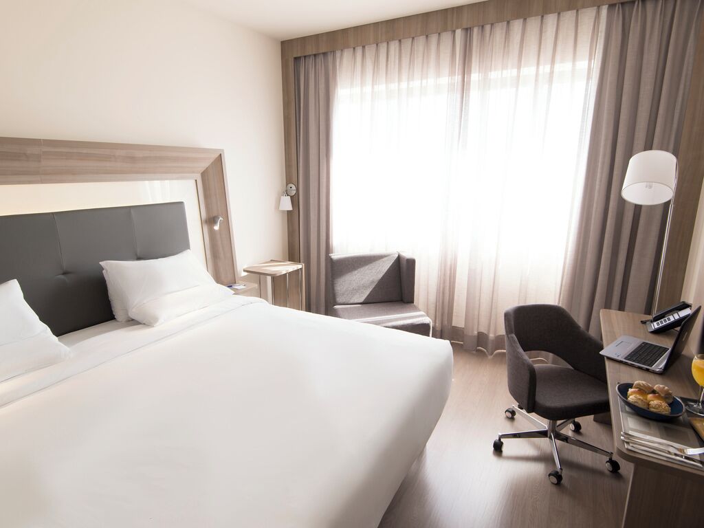 A hotel room with a large bed, a desk with a laptop, a chair, and a small table with a lamp.