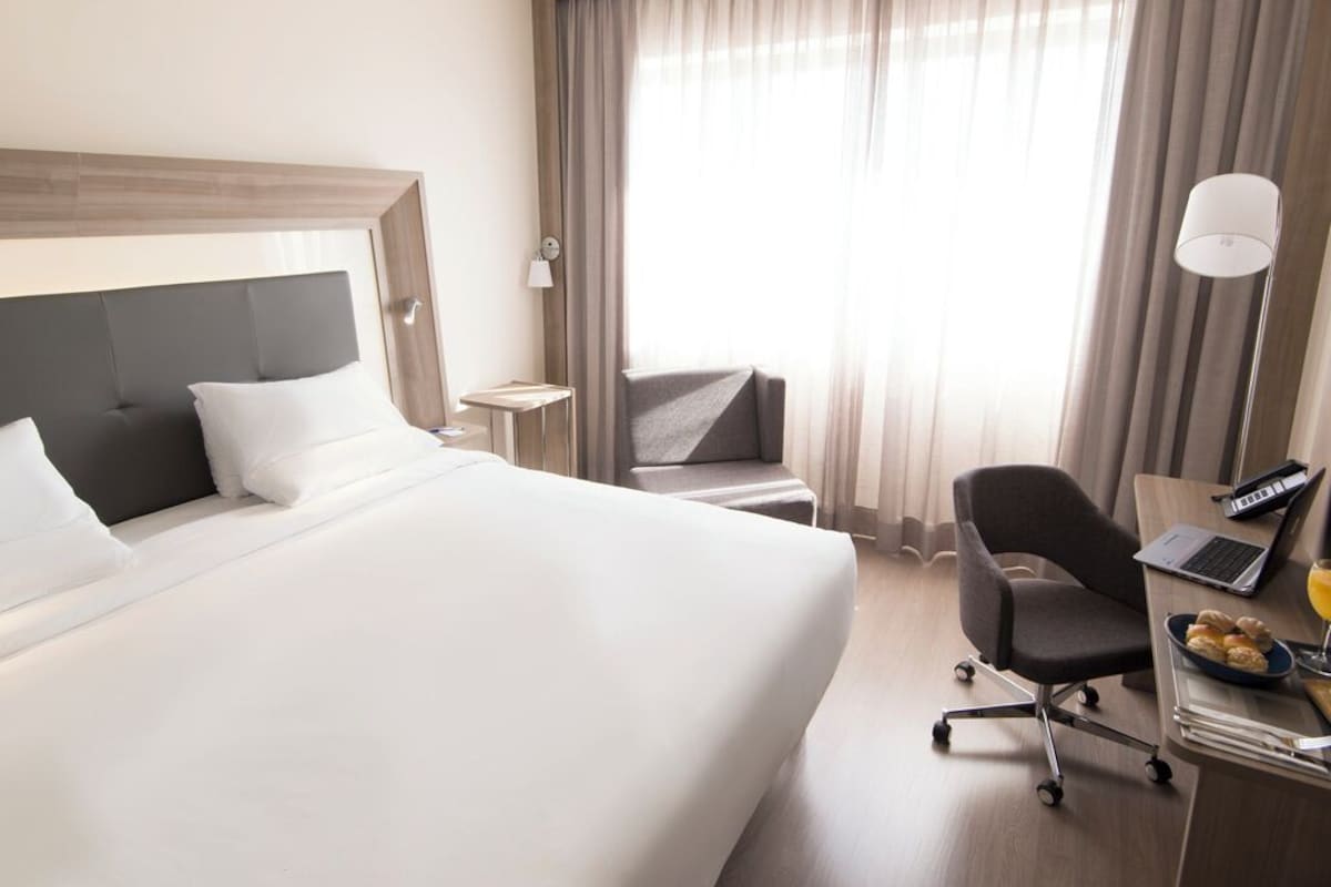 A hotel room with a large bed, a desk with a laptop, a chair, and a small table with a lamp.