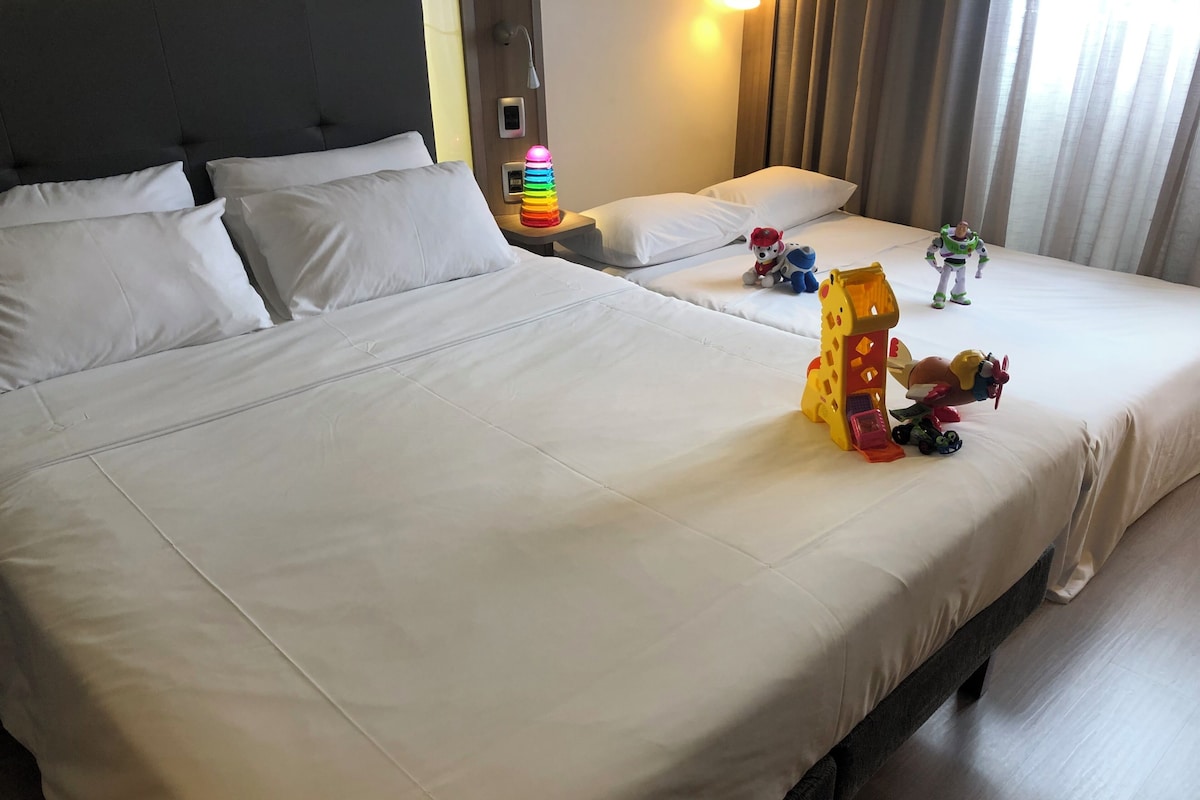A hotel room with a large bed, a bedside table with a lamp, and toys on the bed.
