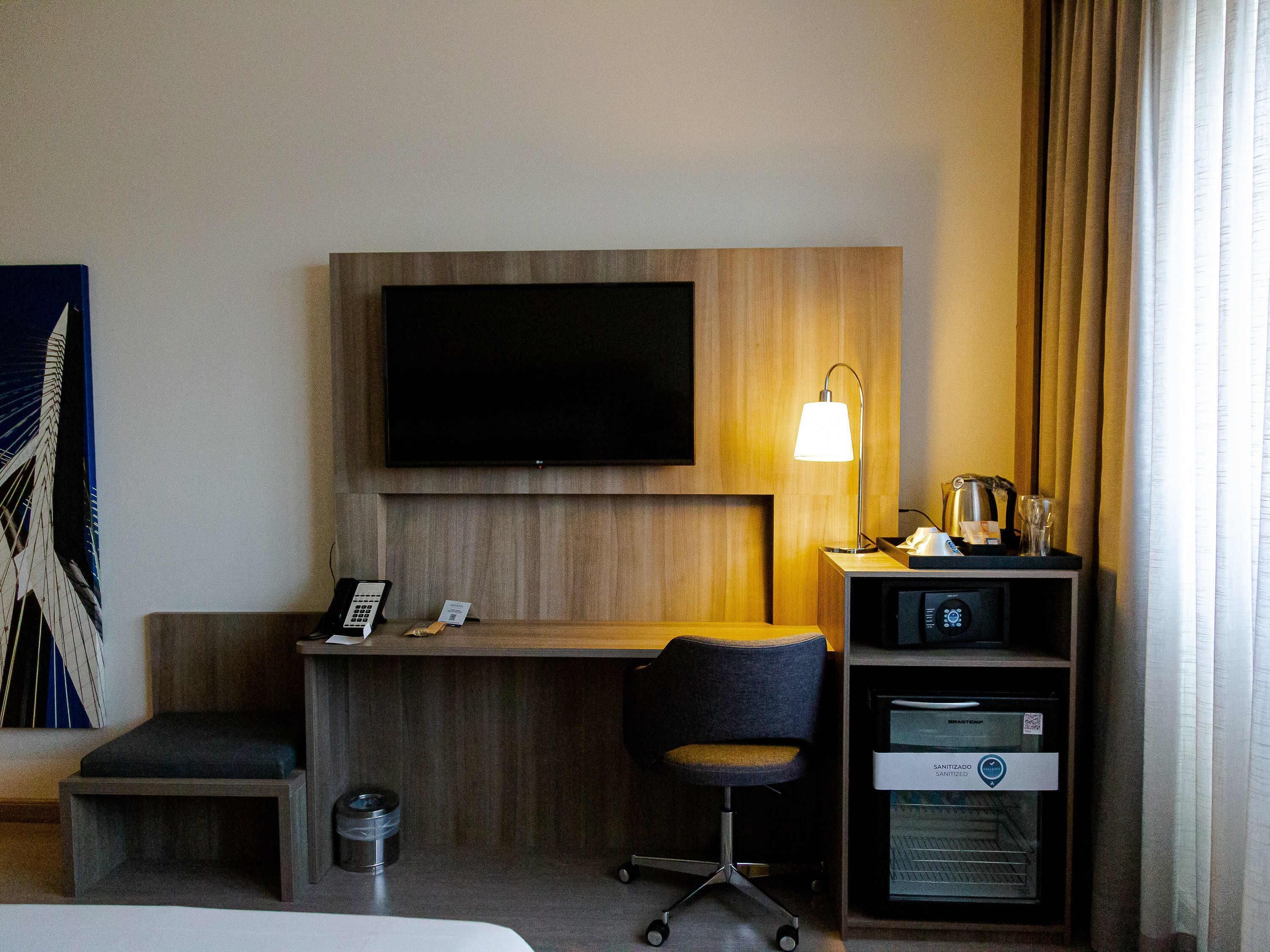 A hotel room with a bed, a desk with a chair, a television, a lamp, a minibar, and a trash bin.