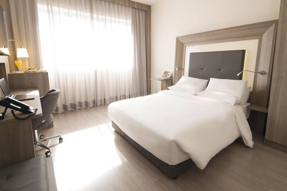 Una camera d'albergo con un letto, una scrivania, una sedia, una lampada e una finestra con tende.