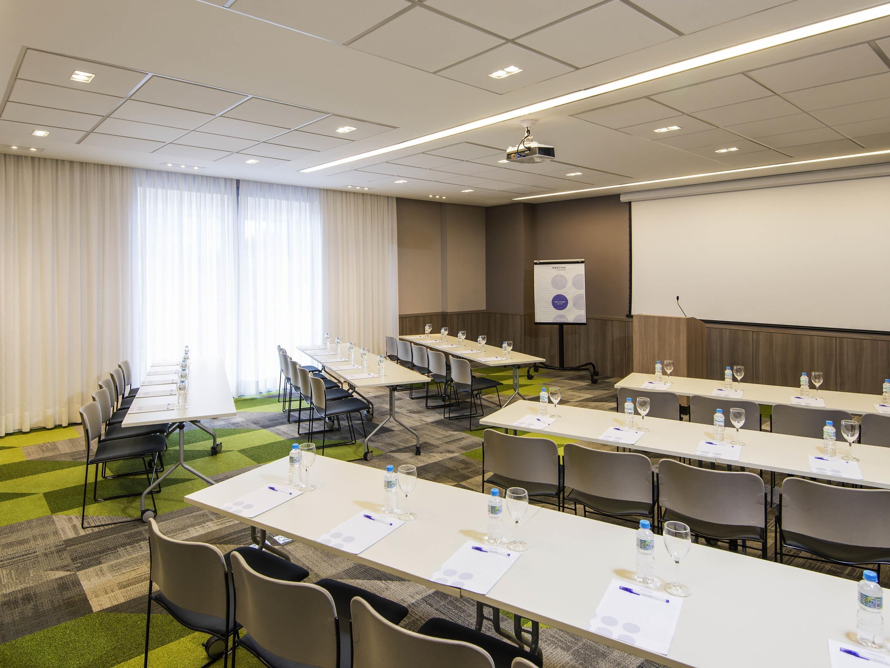 Ein Konferenzraum mit langen Tischen, Stühlen und einer Projektionsleinwand.
