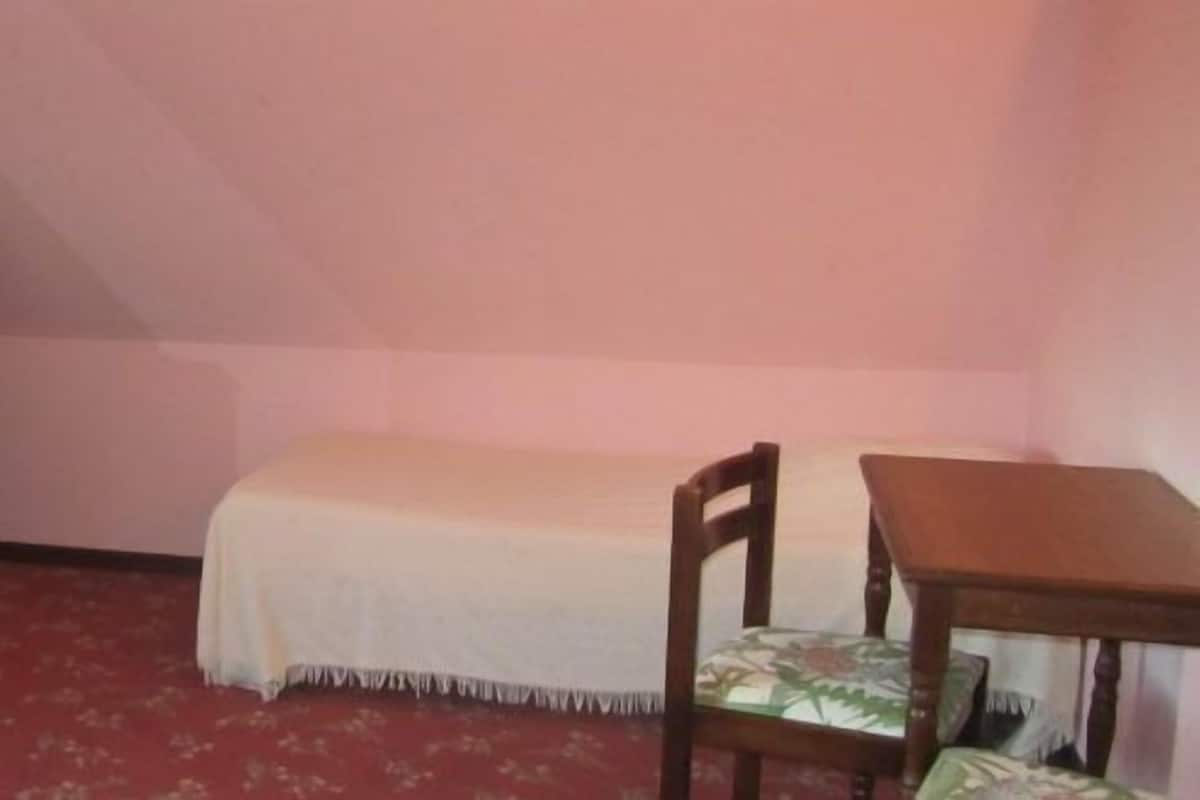A small, dimly lit room with a single bed, a small table, a television on a cabinet, and a chair with a floral cushion.