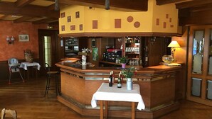 A bar area with a wooden counter, a table with a white tablecloth, and a chair. There are bottles on the counter and a lamp on the right side. - Hotel aux deux clefs (Marmoutier)
