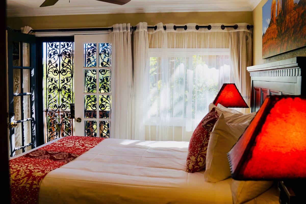 A bedroom with a bed, a window with curtains, a door with a metal gate, and a red lamp.