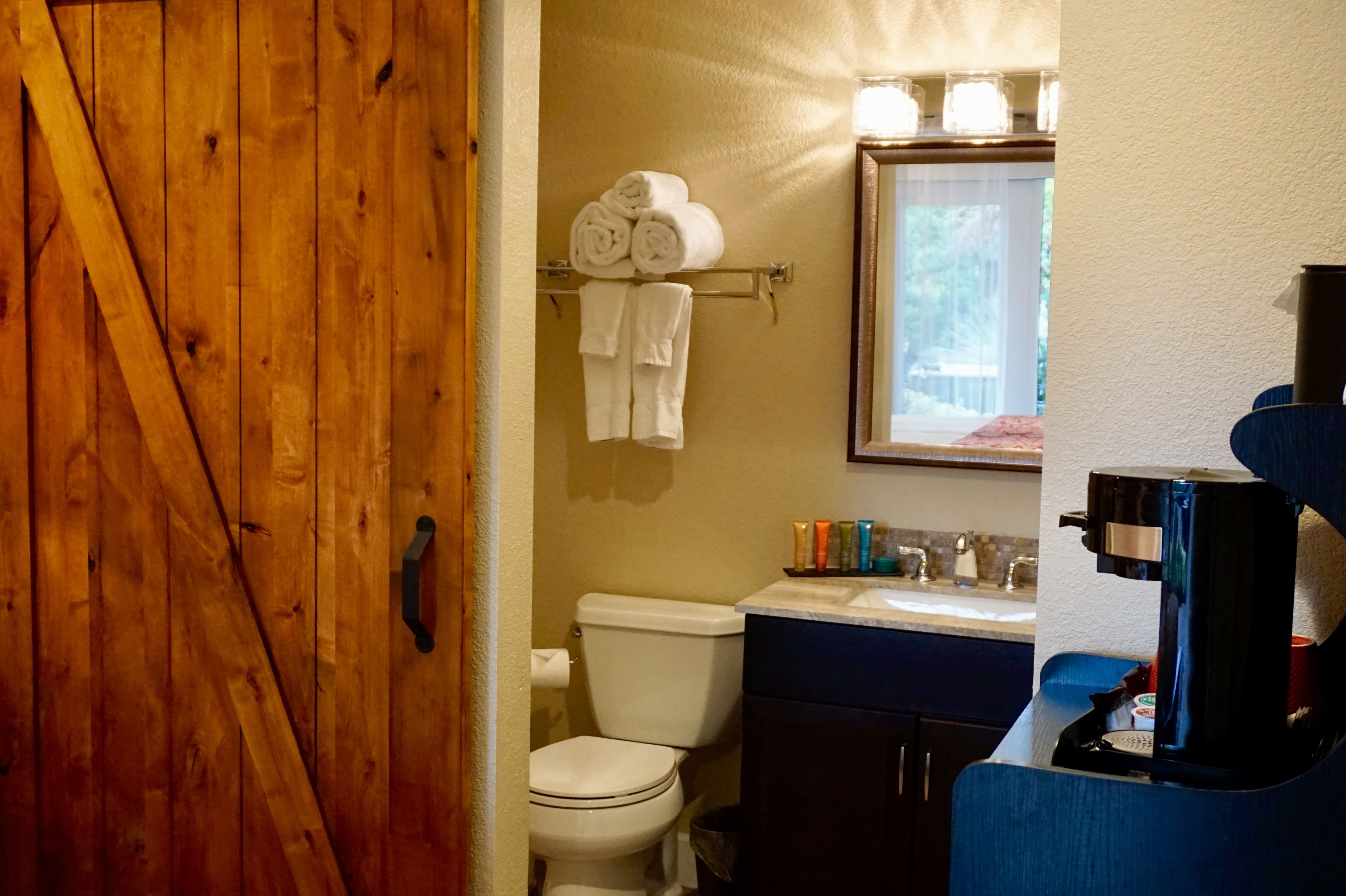 A bathroom with a wooden door, a toilet, a sink with toiletries, and a coffee maker.