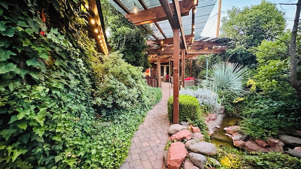 A garden pathway with a pergola overhead, surrounded by lush greenery and a small water feature.