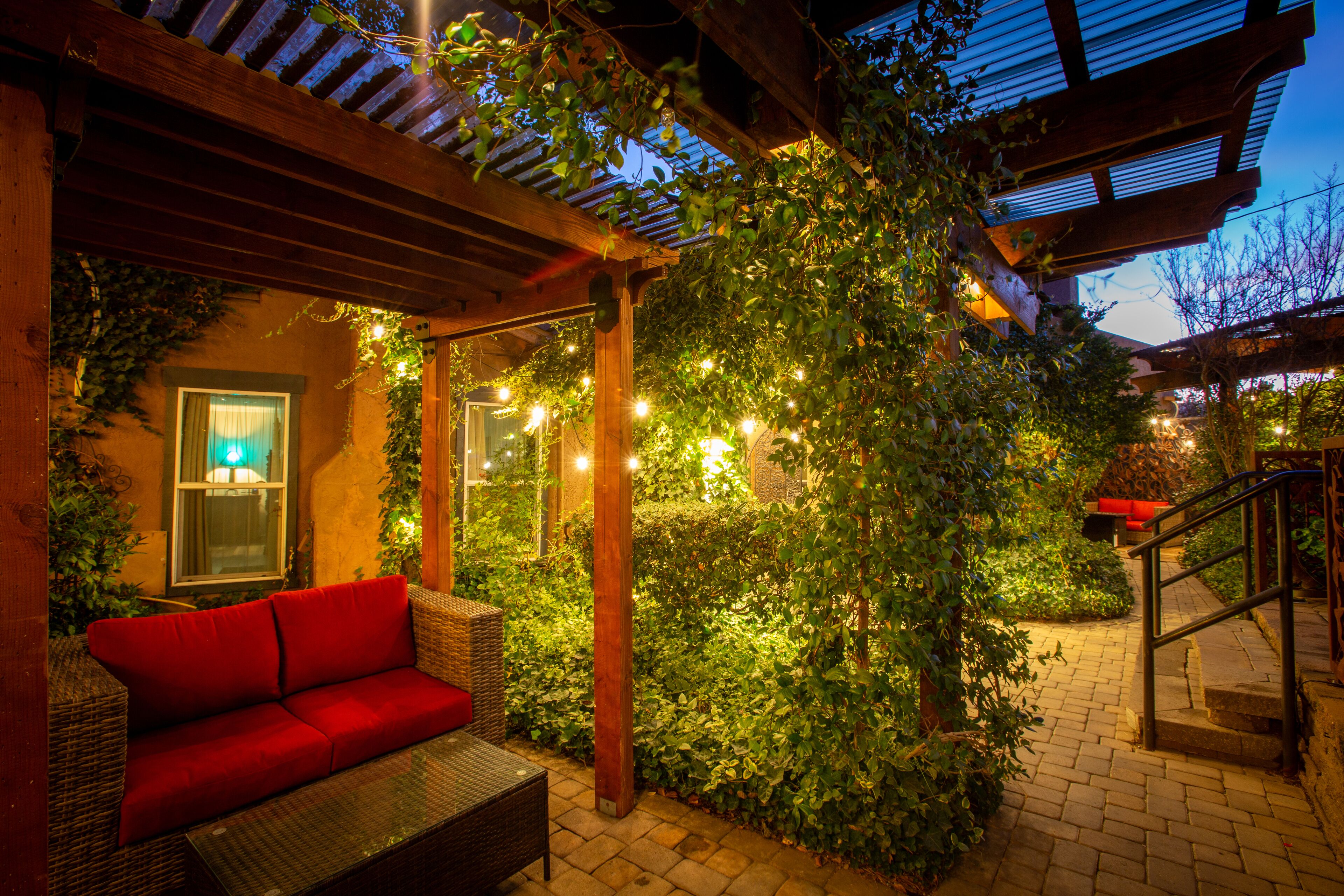 A covered outdoor seating area with a red sofa and wicker furniture, surrounded by greenery and string lights.