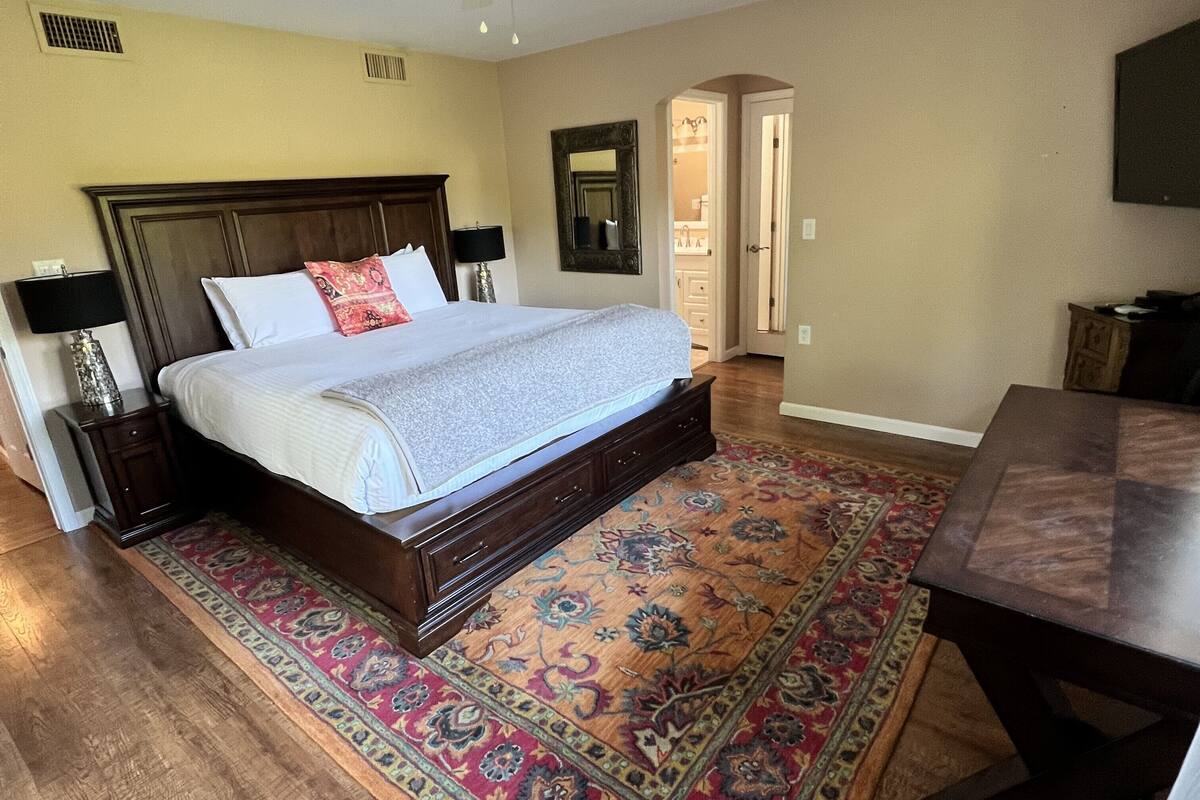 A bedroom with a large bed, wooden headboard, two bedside tables with lamps, a patterned rug, and a wooden nightstand.