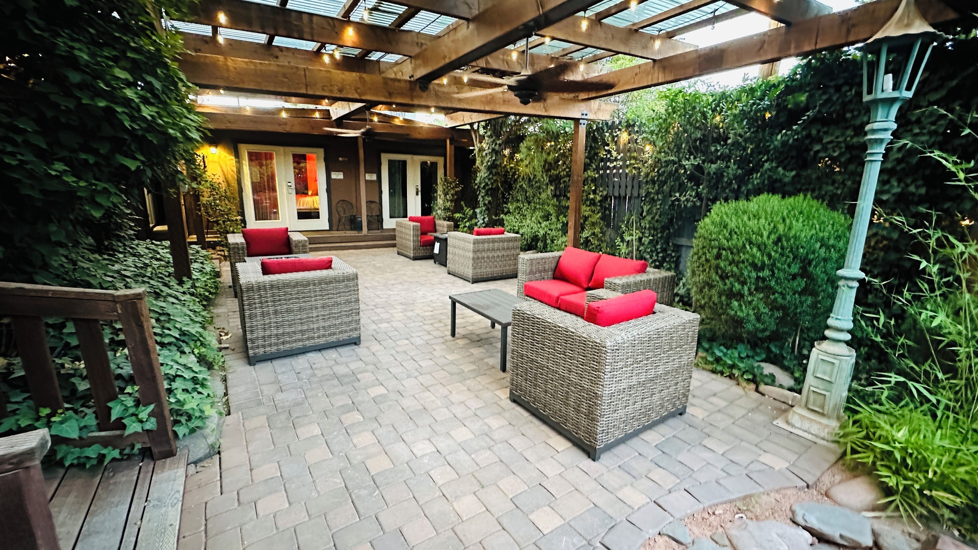 An outdoor patio area with wicker furniture and red cushions, surrounded by greenery and a wooden pergola with string lights.