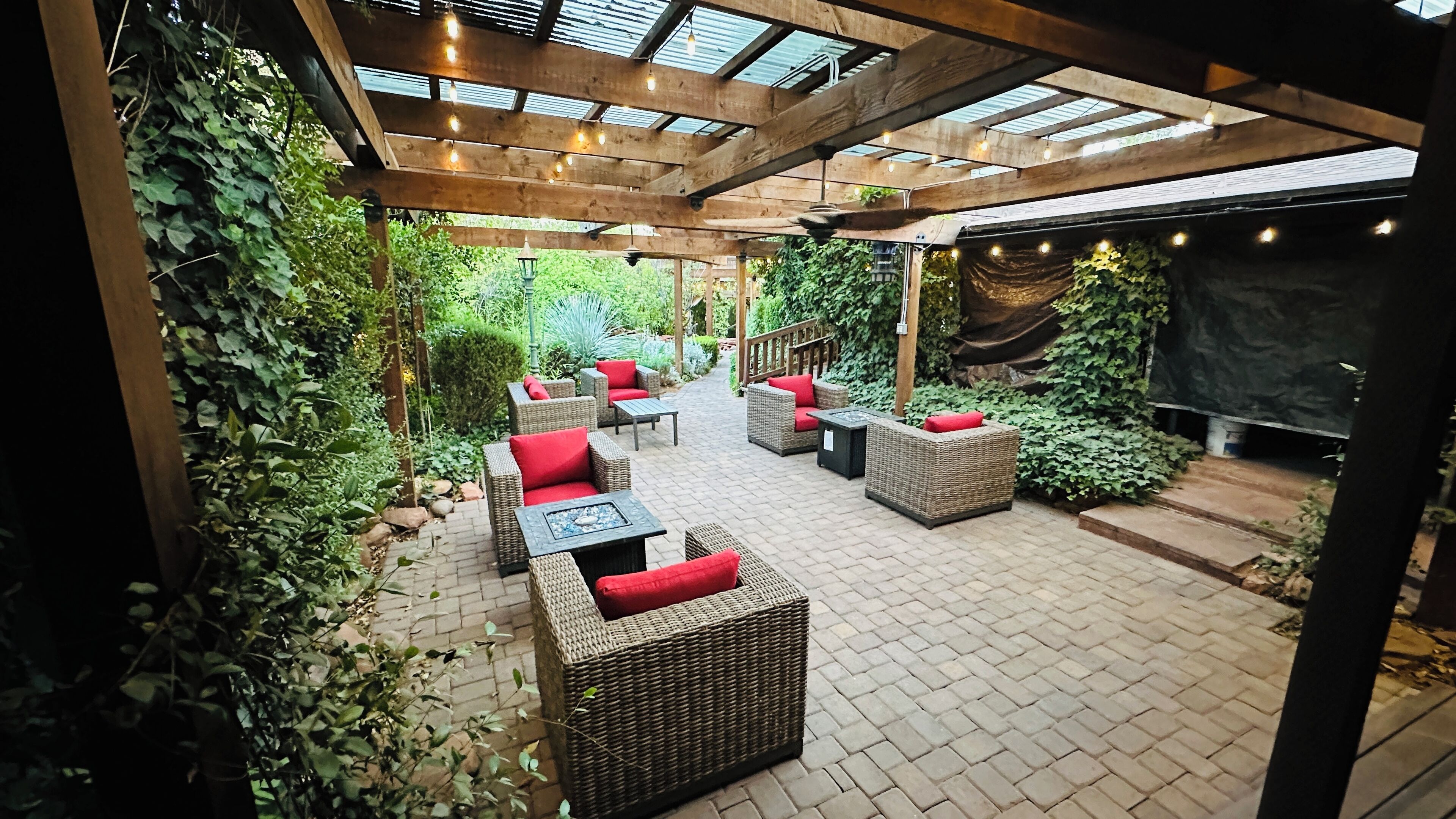 An outdoor patio with wicker furniture and red cushions, surrounded by lush greenery and a brick floor.