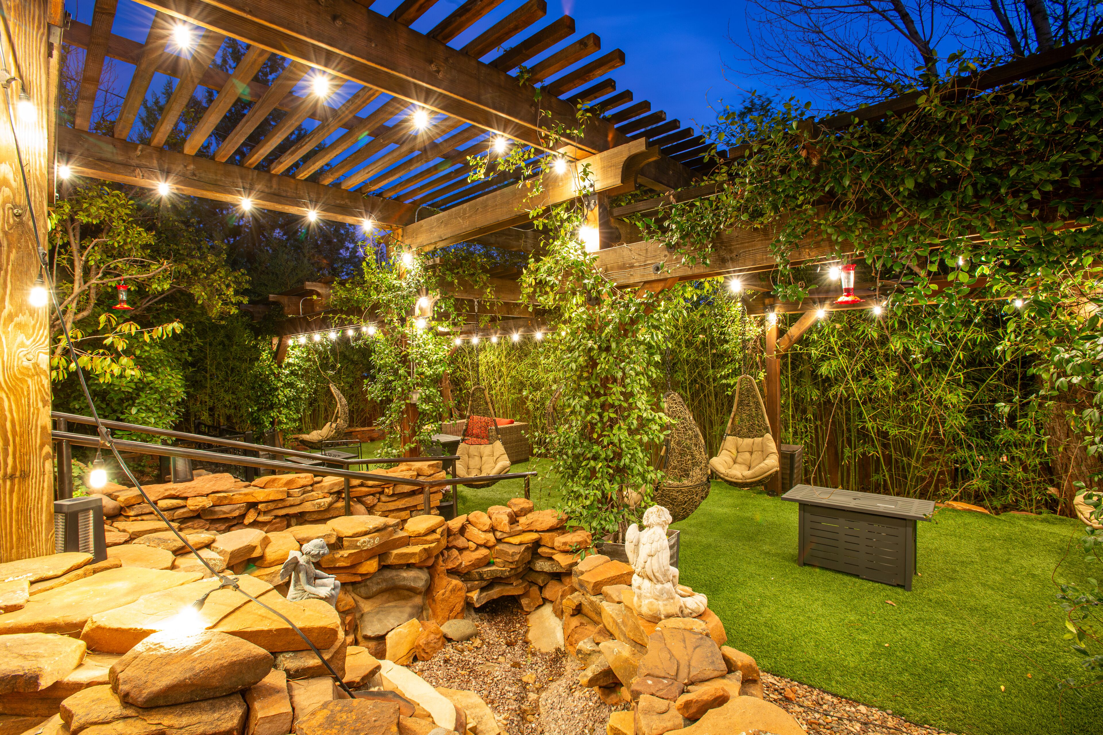 Een tuin met een pergola, lichtjes, een rotsmuur en een beeld.