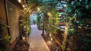 A brick pathway flanked by greenery and string lights, leading to a building entrance.