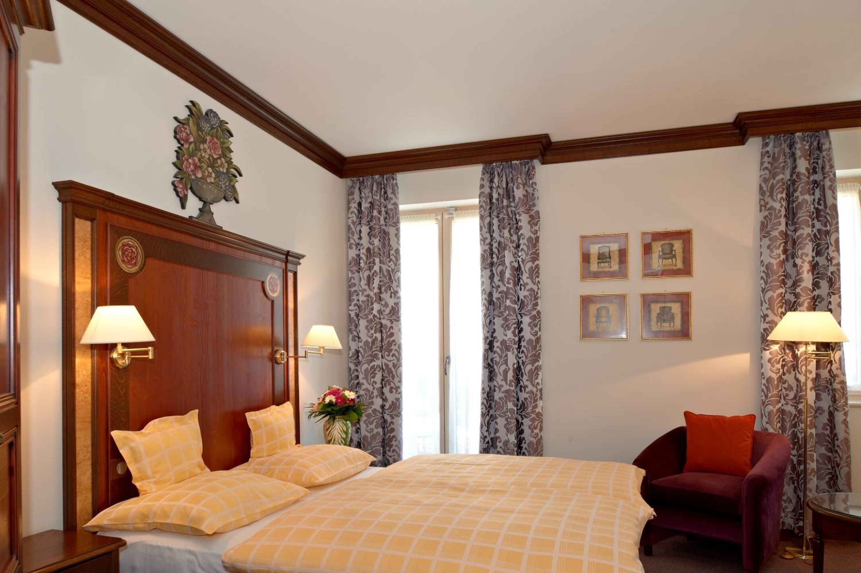 Une chambre d’hôtel avec un grand lit, deux lampes de chevet, une tête de lit en bois, un fauteuil rouge et une fenêtre avec des rideaux.