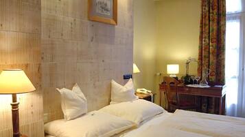 A hotel room with a large bed, two bedside lamps, a wooden headboard, a framed picture, a desk with a telephone, and a chair.