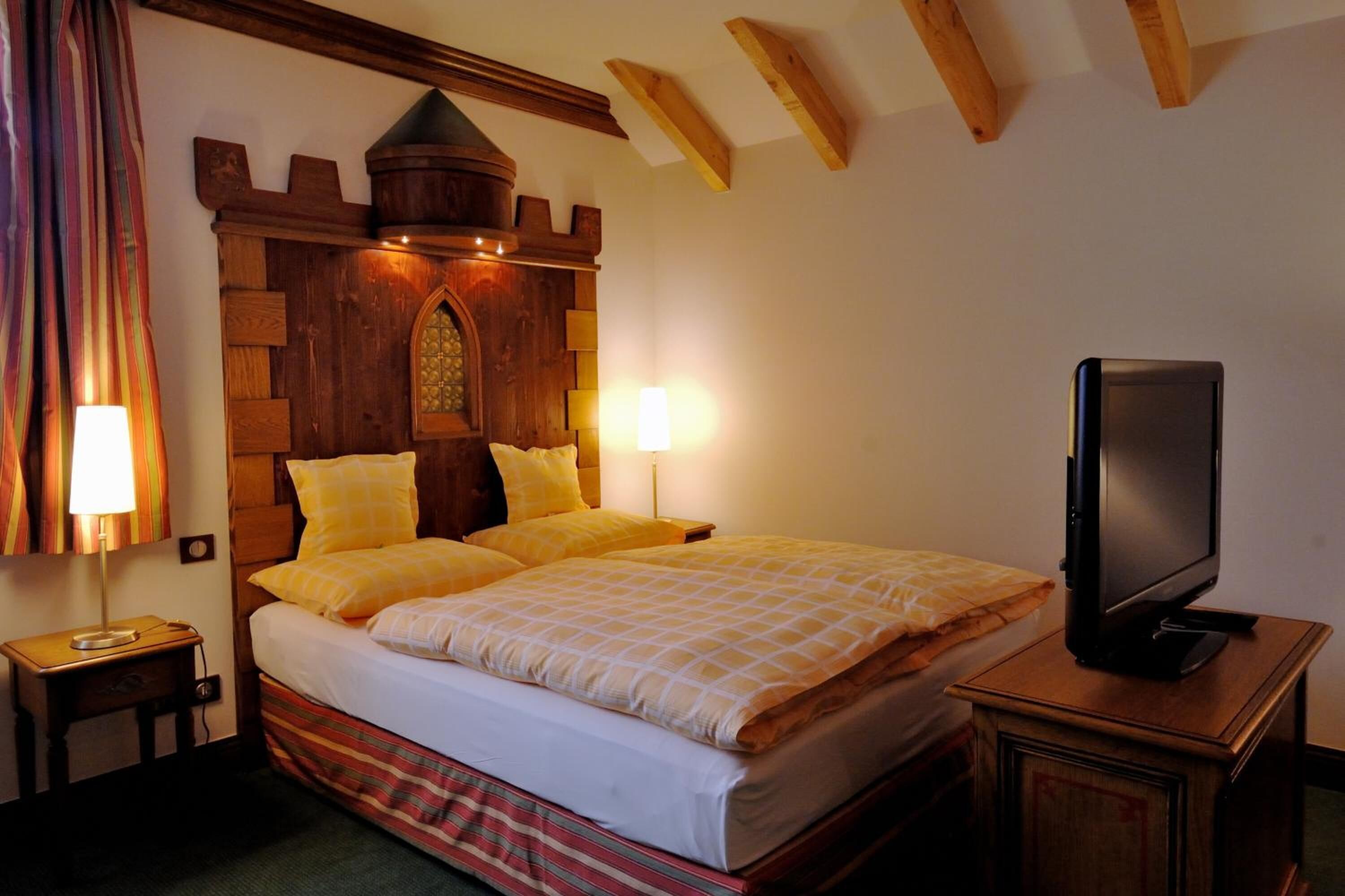 Un dormitorio con cabecera de madera, dos lámparas de noche, un televisor sobre un mueble de madera y una cama con ropa de cama amarilla.