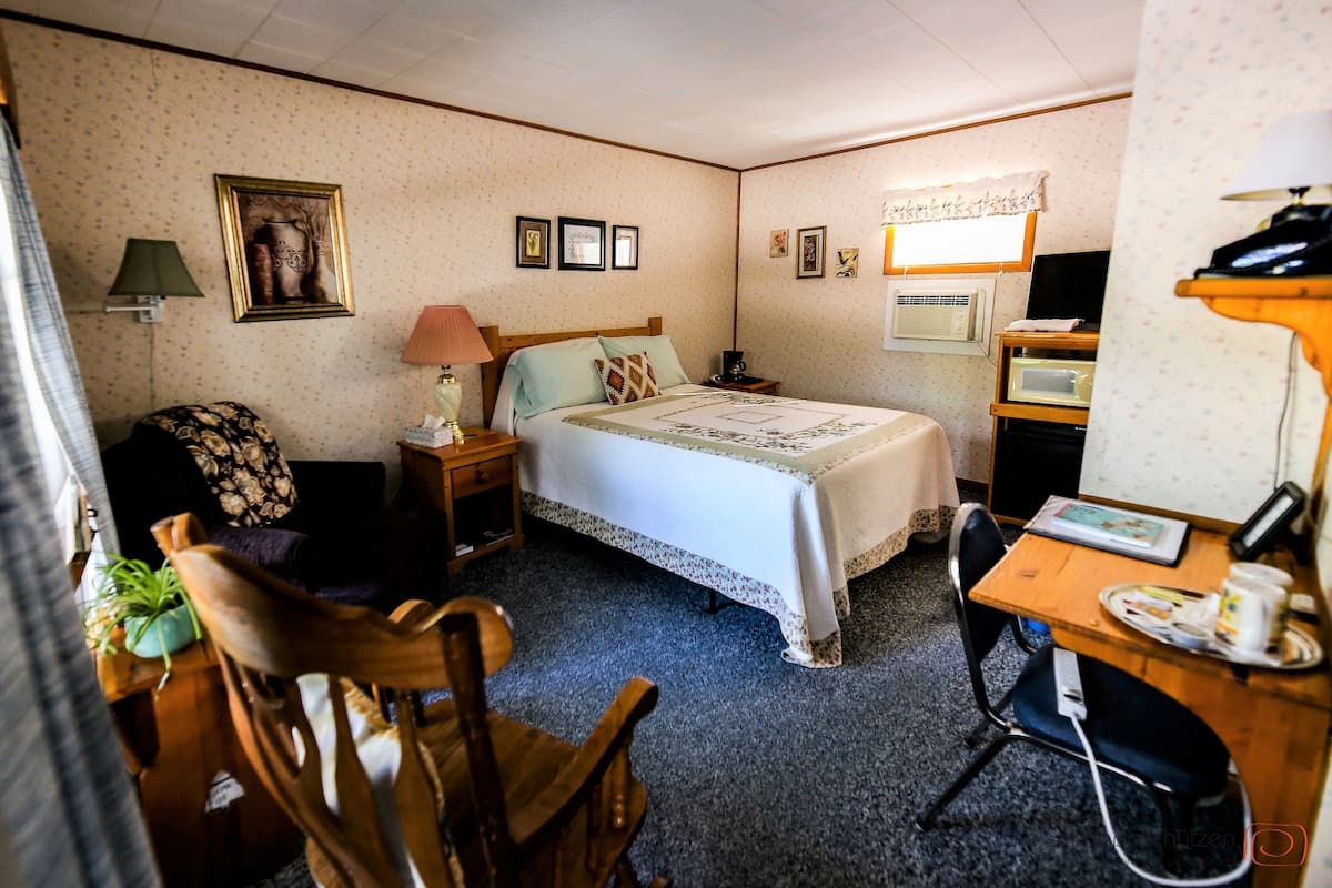 A small, cozy room with a bed, a desk, a chair, a lamp, a television, and a wall-mounted air conditioner.