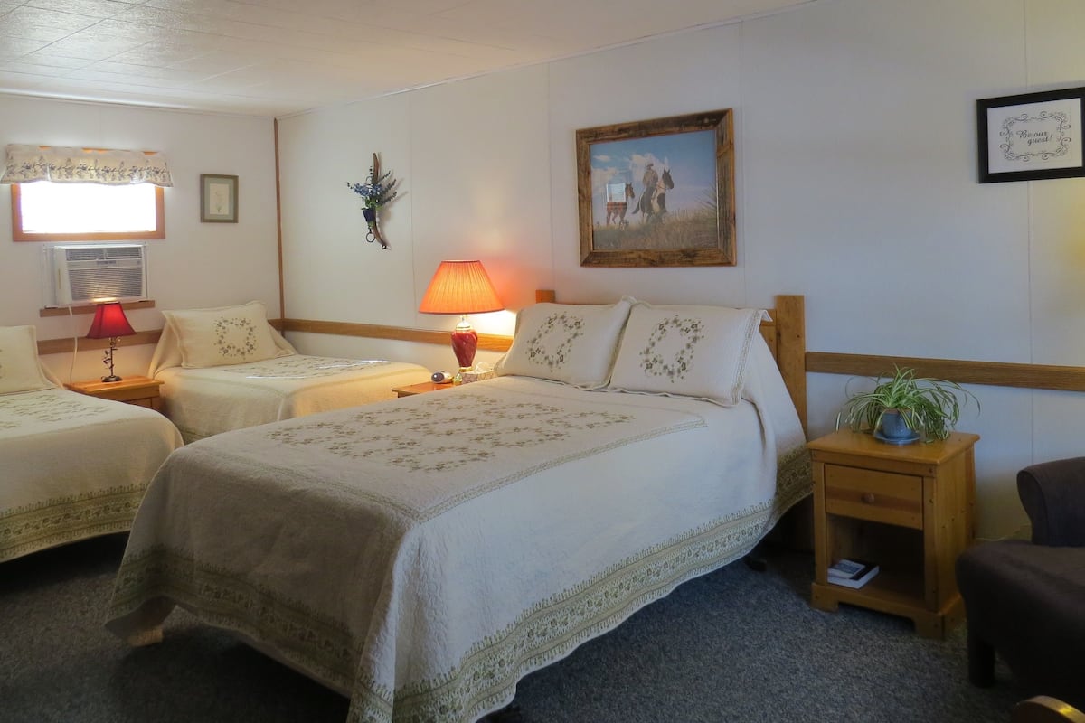 A hotel room with two beds, a wooden headboard, a painting, a wall-mounted air conditioner, and a small table with a lamp.