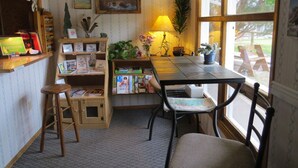 Une pièce chaleureuse avec une table en bois, des chaises, une bibliothèque remplie de livres et de cartes, un tableau encadré, une lampe et une fenêtre donnant sur l’extérieur.