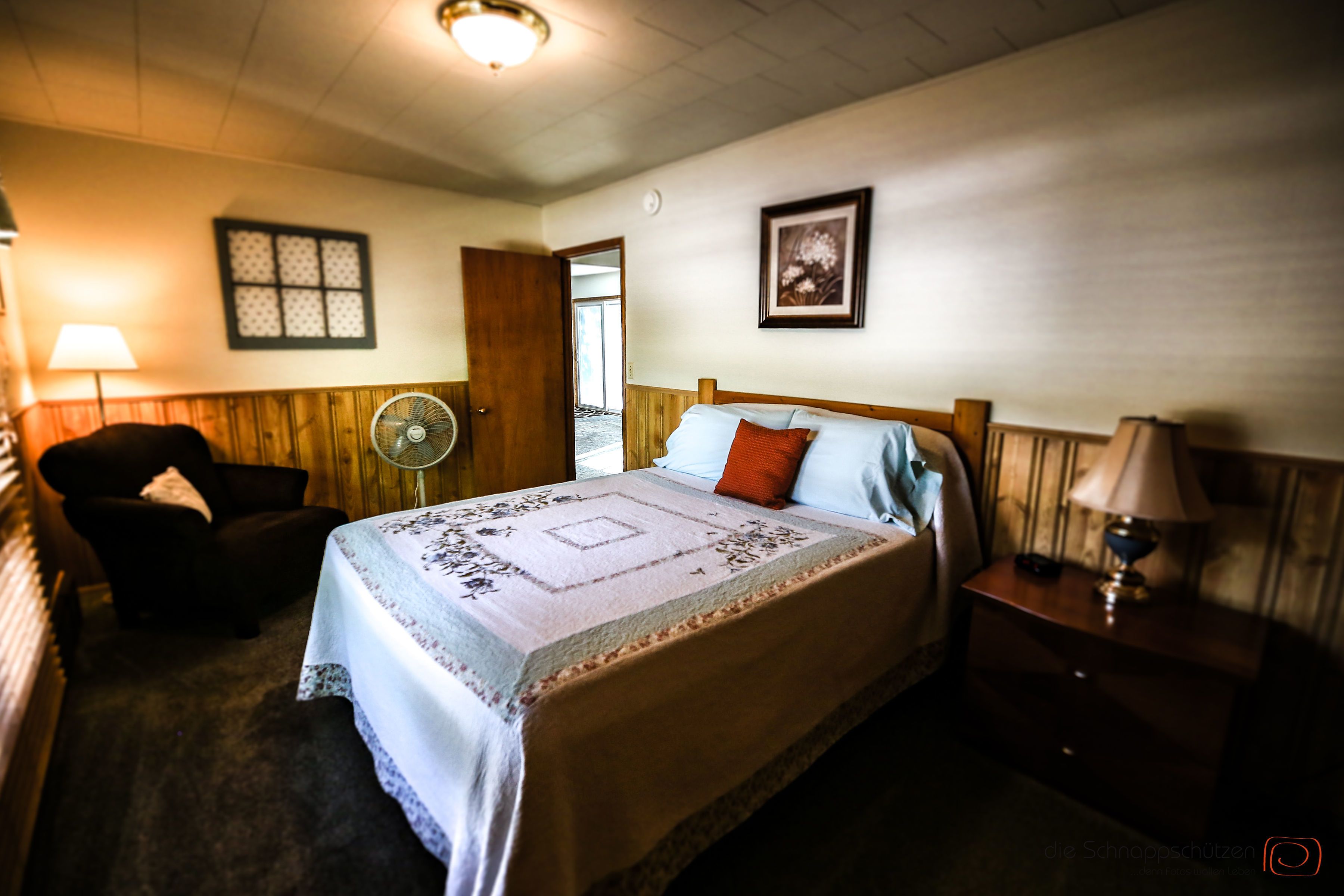 A bedroom with a bed, bedside table, lamp, chair, and a fan.