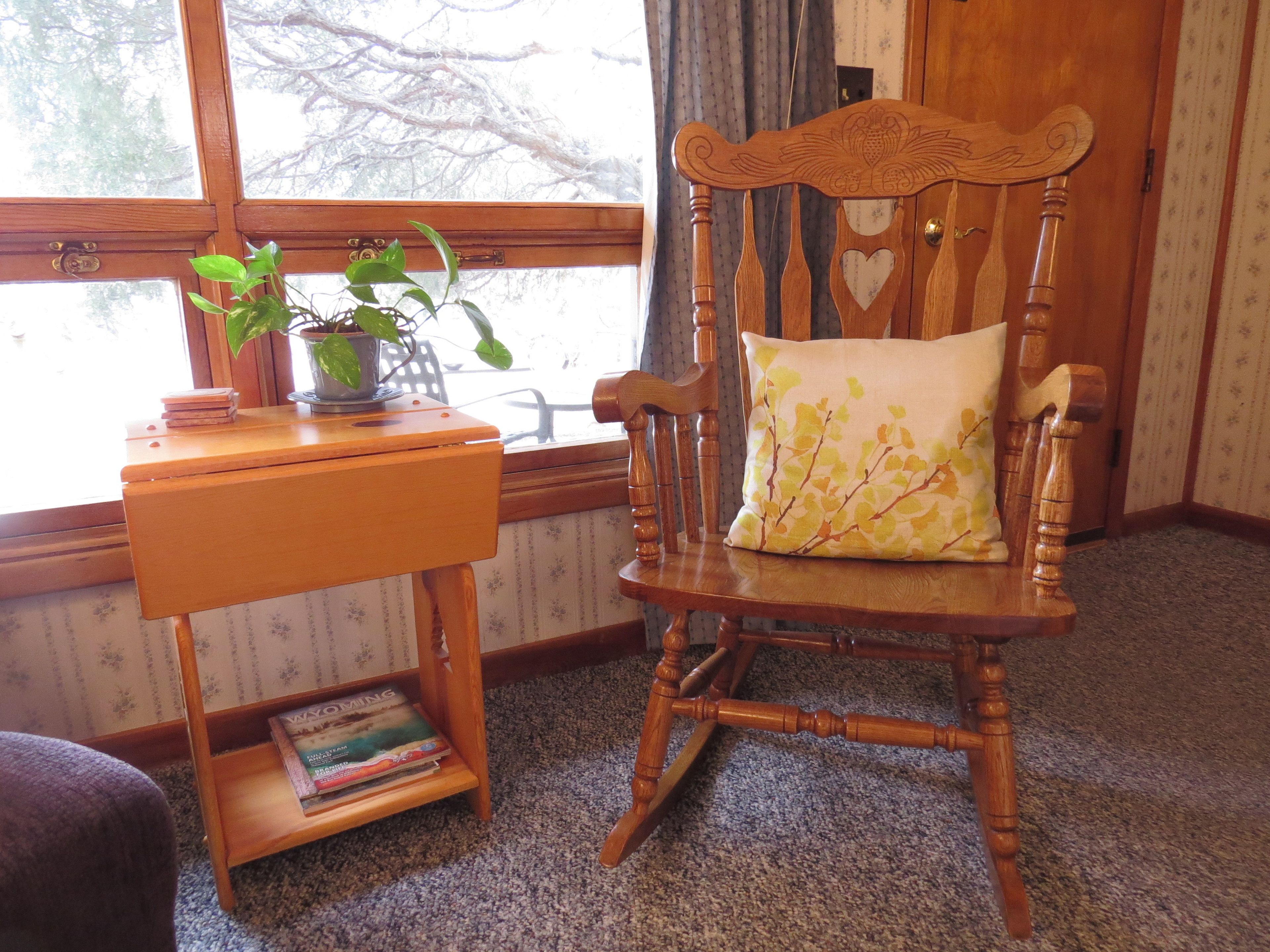 A wooden chair with a cushion, a small wooden table with a plant, and a window with a view of trees.