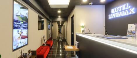 A hotel reception area with a modern design, featuring a reception desk, a red seating area, and a large digital display on the wall.