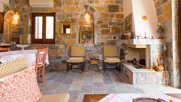 Una habitación con suelo de piedra, una chimenea, dos sillas de madera, una mesa con mantel blanco y una cama con ropa de cama estampada.