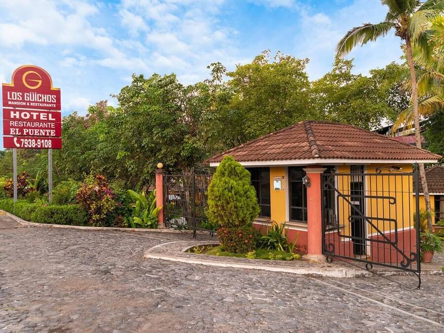 Un pequeño edificio de una sola planta con tejado de tejas, rodeado de exuberante vegetación y un sendero adoquinado.