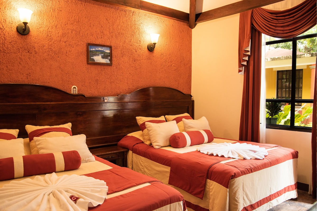 Habitación de hotel con dos camas, cada una con ropa de cama roja y blanca, cabeceros de madera y dos lámparas de pared.