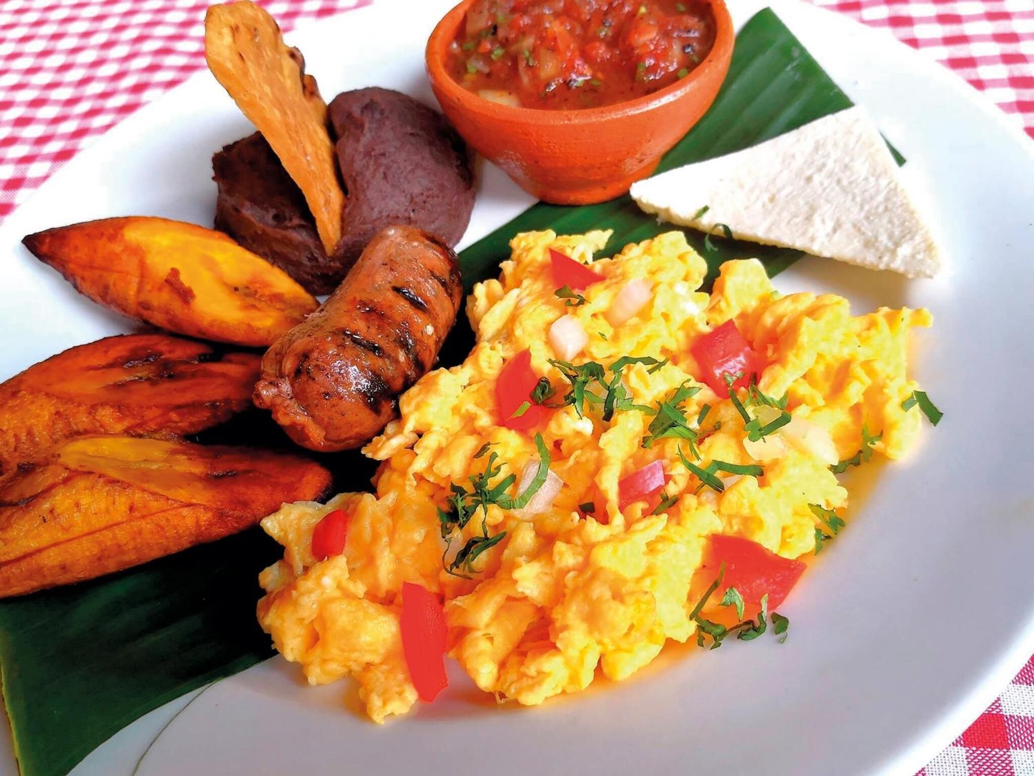 Un plato de comida con huevos revueltos, plátanos maduros, salchichas y un tazón de salsa.