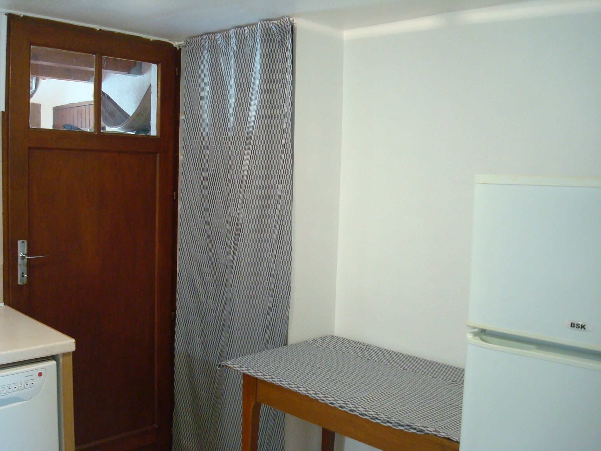 A small, compact kitchen with a refrigerator, a wooden door, and a window.