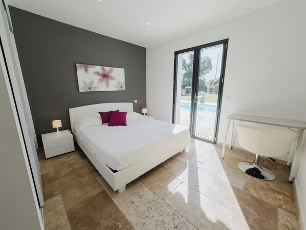 A modern bedroom with a large bed, a nightstand, a desk, and a view of a swimming pool through a sliding glass door. - STE LUCIE DE PORTO VECCHIO villa / heated pool (classified ****) (Zonza)