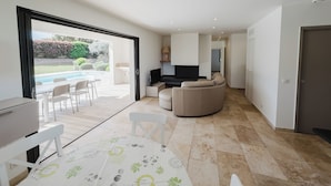 A modern living room with a sofa, a TV, and a dining area with a table and chairs. There is a sliding glass door leading to a patio with a pool.