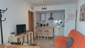 Una sala moderna con una mesa de comedor de madera, un sofá y un televisor. Se ve una cocina al fondo con un microondas, un lavabo y taburetes.