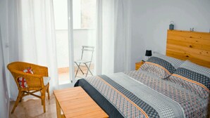 Un dormitorio con cama, cabecera de madera, una silla de mimbre, una mesita y una ventana con cortinas.