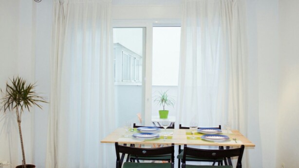 Un comedor con una mesa dispuesta para dos, una planta en maceta y una ventana con cortinas.