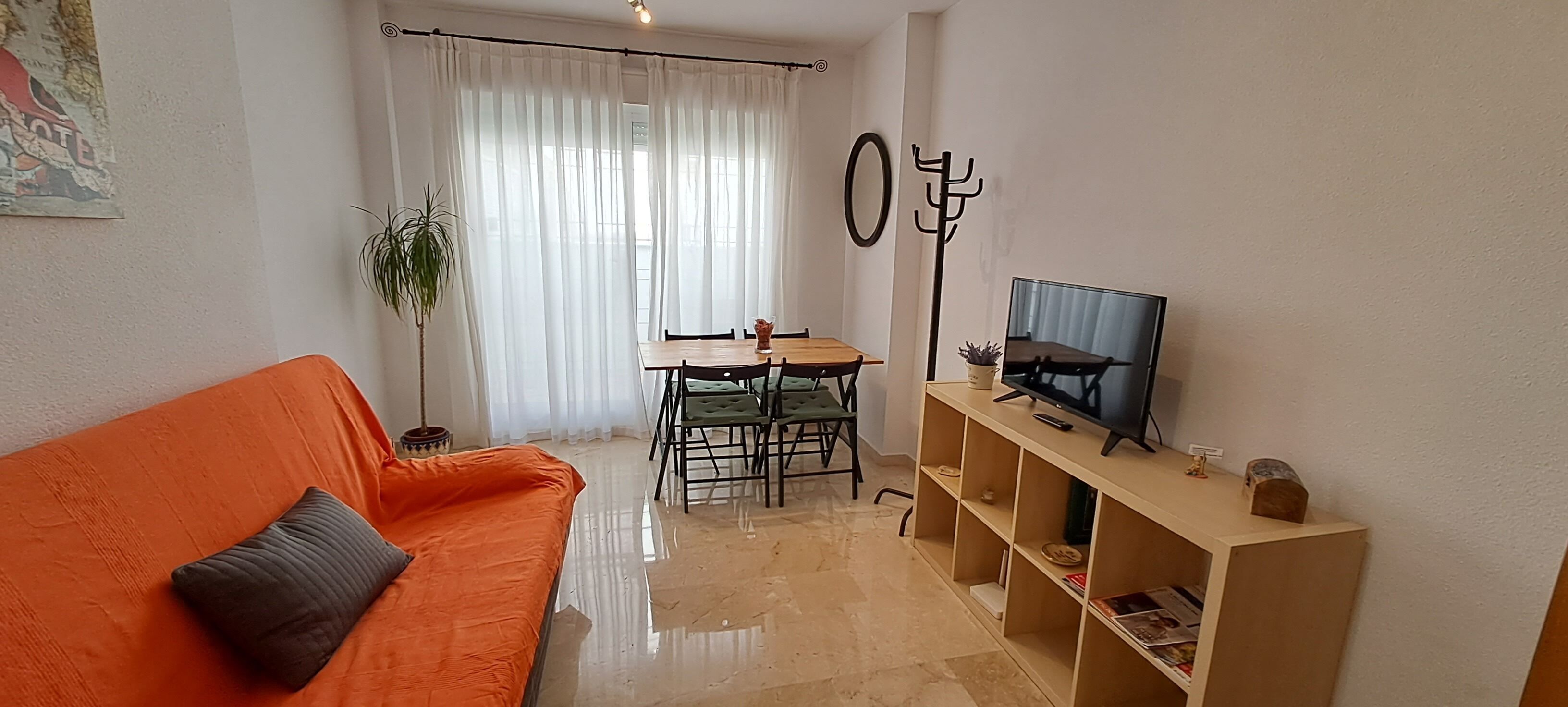 Uma sala de estar com sofá, uma mesa de jantar com cadeiras, uma televisão sobre um móvel e um vaso de planta.