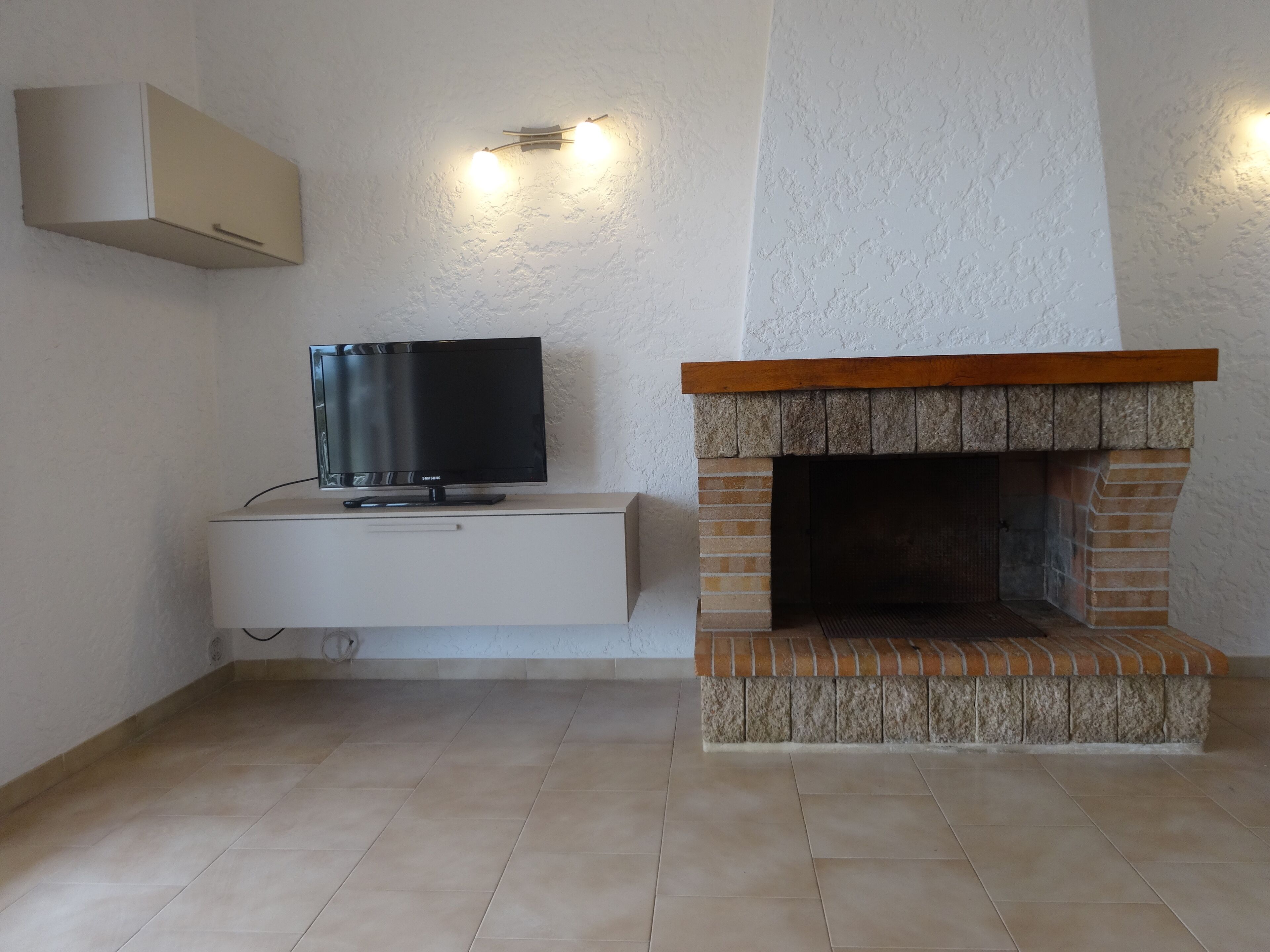 A room with a flat-screen TV on a white cabinet, a stone fireplace, and a wall-mounted light fixture.
